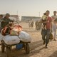 Photograph showing crowd of migrants walking with carts of their belongings; man holding small child