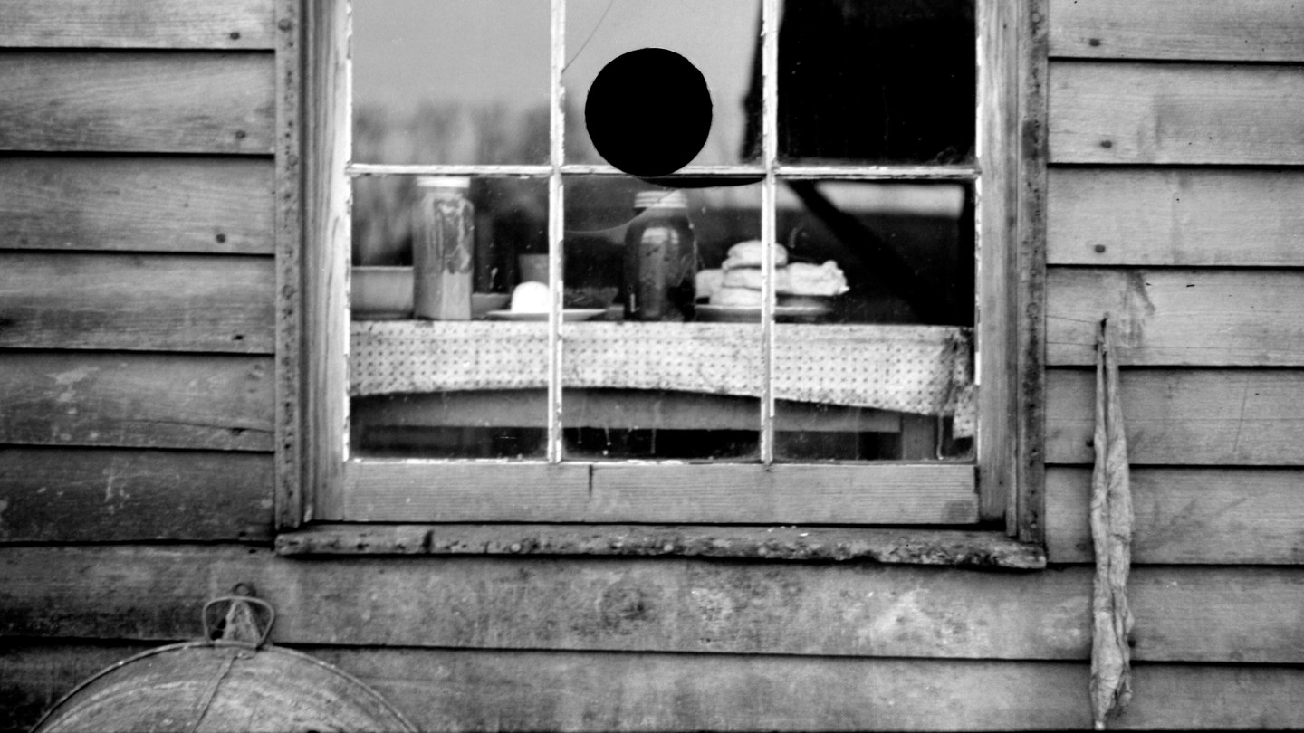 A black and white photo of a window in a wooden house, showing a table inside with jars on it