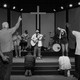 A black-and-white photo of a group standing in a church, listening to someone at the front playing a guitar