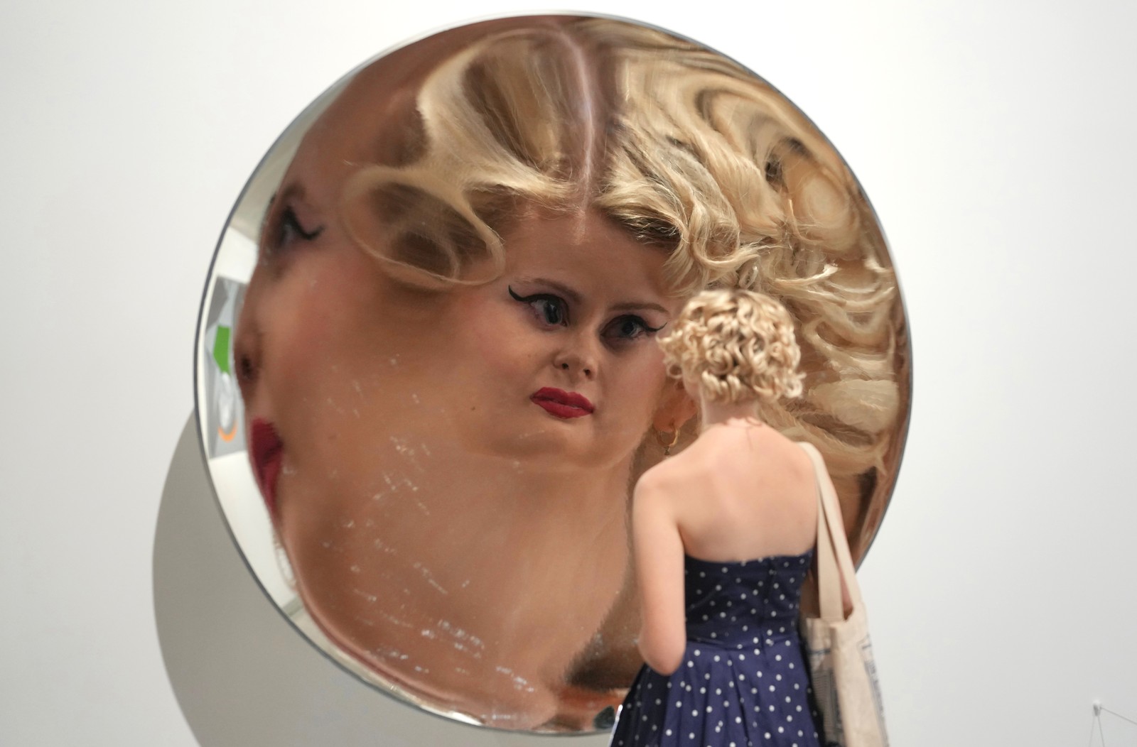 A person stands close to a huge circular curved mirror, their face magnified and distorted in the reflection.
