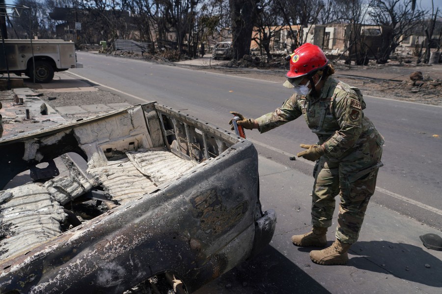 A person wearing a military uniform and a hard hat spray-paints a symbol on the back of a burned pickup truck.