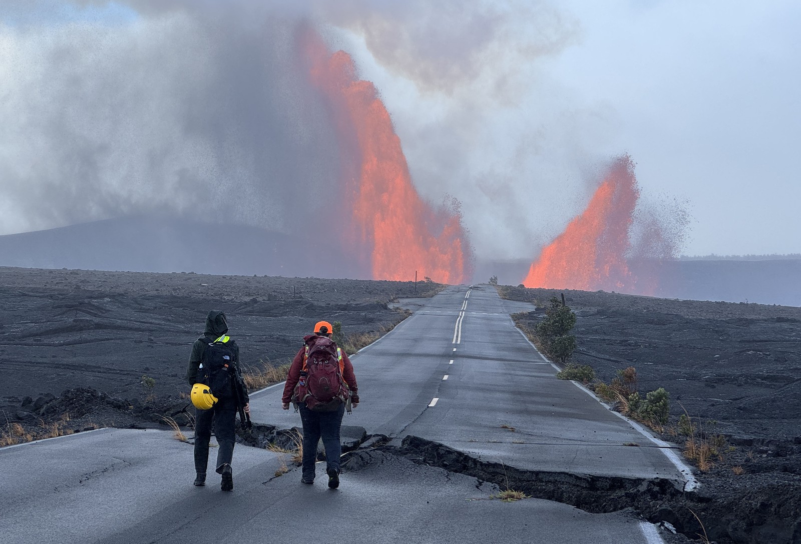 Two people stand on a fractured and damaged road, looking toward a double fountain of lava erupting ahead of them.
