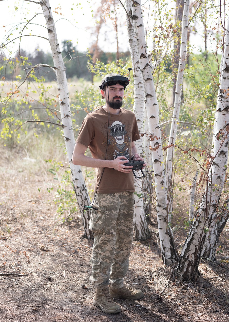 A thin, beaerded man in military fatigues and an animated t-shirt holding a drone operating controller.