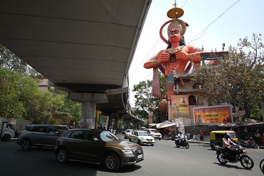 Cars and motorcycles drive on a road past an overpass and a large statue of a Hindu god.