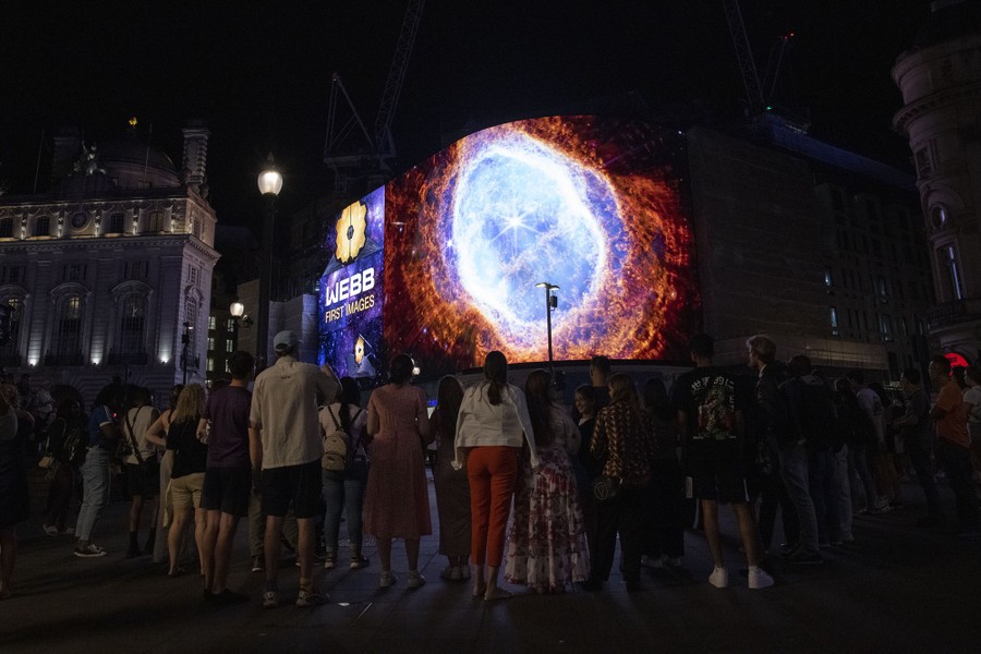 A crowd gathers in a city square, looking up at a large display screen showing images of deep space.
