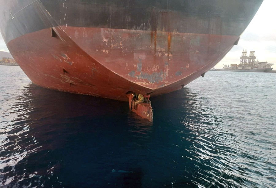 Three people sit atop the rudder of a large tanker, just above the water's surface.
