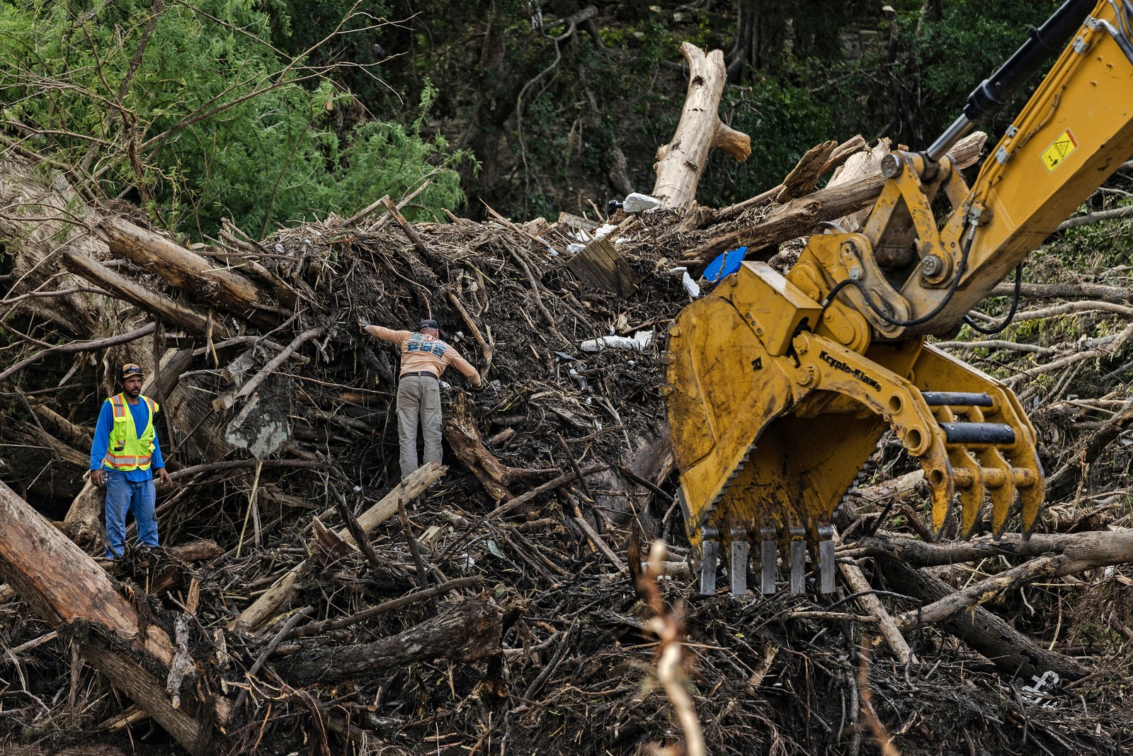 Search and recovery workers stand on piles of debris as an excavator works to clear flood damage.