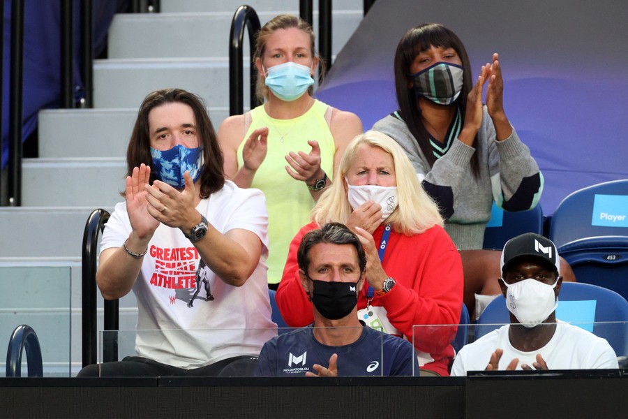 A group of six masked people sit together in the stands, applauding.