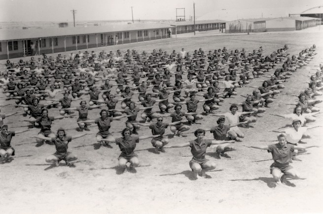 black-and-white archival photo of dozens of women exercising in rows on field, squatting with arms stretched out 