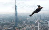 A person wearing a parachute in a pack strikes a casual pose in mid-air as they leap from the top of a tall building.