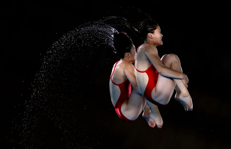 Two synchronized divers mid-jump, holding their knees to their chests while spinning and throwing off water droplets