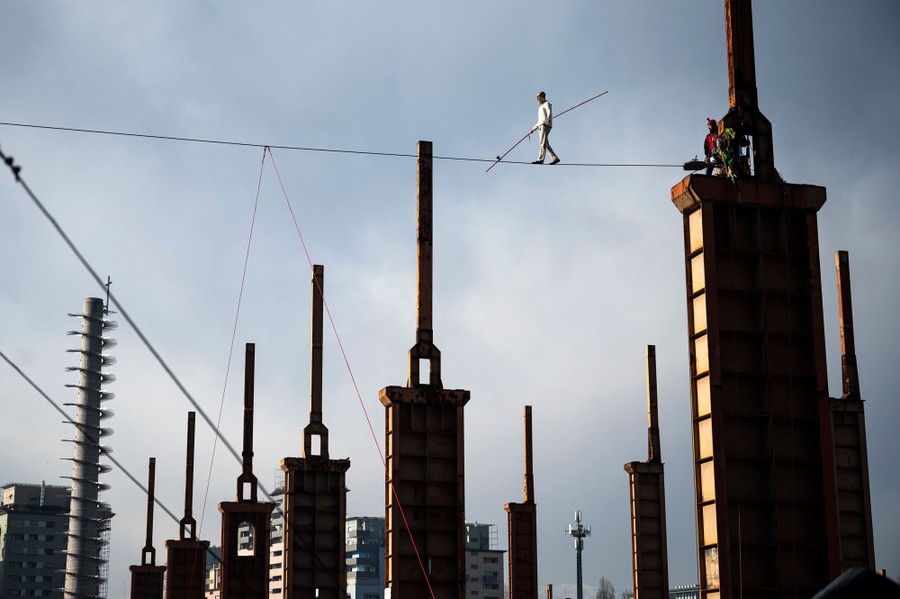 A man holding a long pole walks on a tightrope high above factory towers.