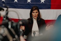 Photo of Nikki Haley speaking at a microphone, hands apart, with press before her and an American flag behind her