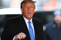 A photo of Donald Trump wearing a black suit and a blue tie, standing with his mouth open as if speaking