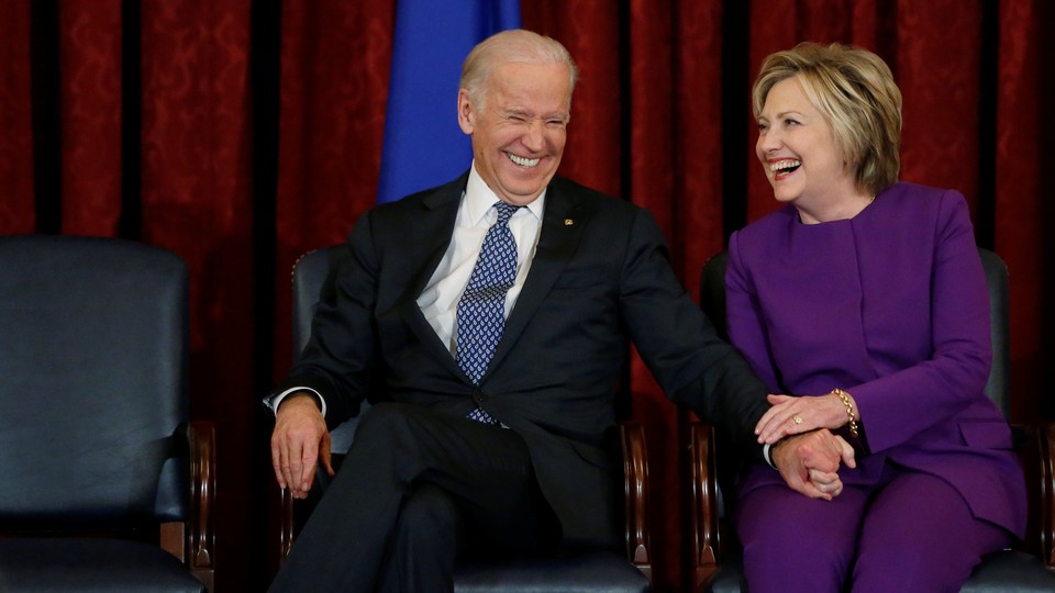 Joe Biden and Hillary Clinton sit on chairs laughing together in front of a red curtain.