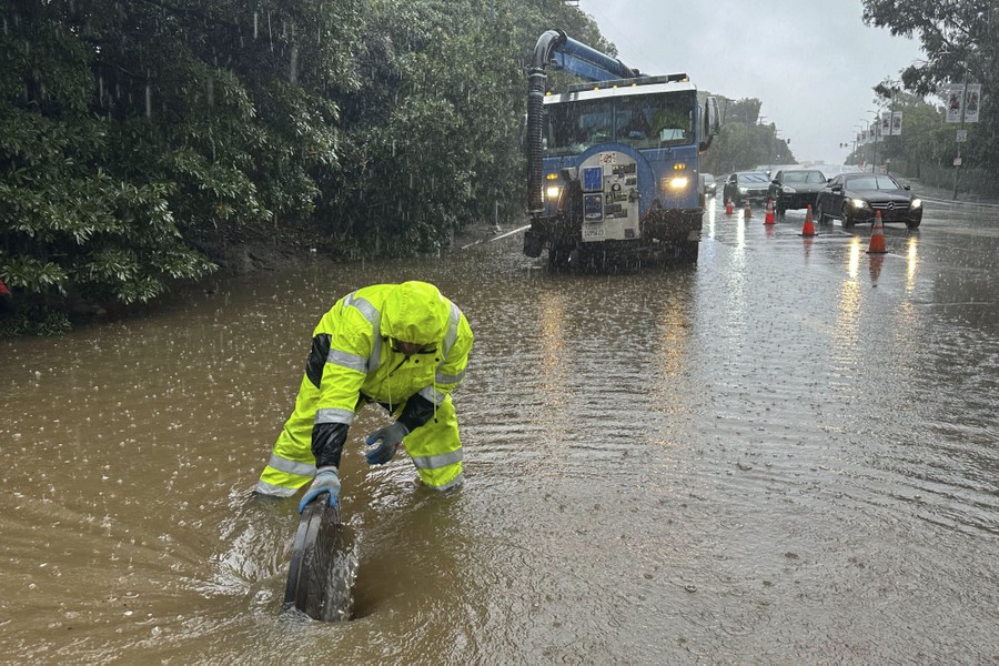 A whirlpool forms around a worker as he lifts a manhole cover to clear knee-deep street flooding.