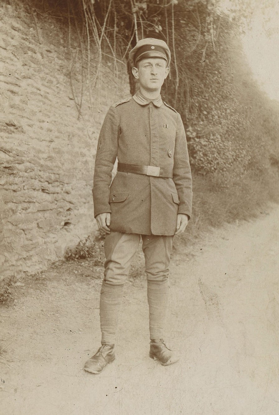 sepia-toned archival photo of uniformed WWI German soldier standing on a road with wall behind
