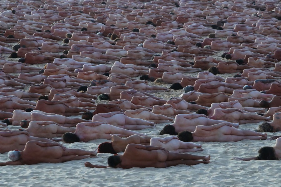 Hundreds of naked people lie on their left side on a sandy beach, arranged for a large-scale photograph.