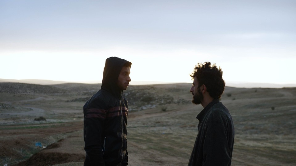 Two men, one in a hoodie and one with shaggy hair, stand in profile in a desert landscape.