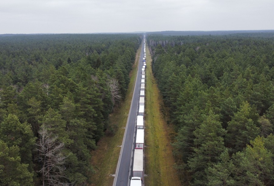 A long line of trucks is seen from above, sitting on a straight road through a forest.