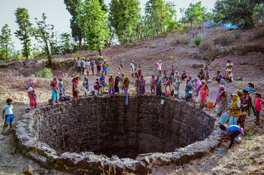 A couple dozen people stand along the edge of a deep and wide well, lowering containers tied to ropes.