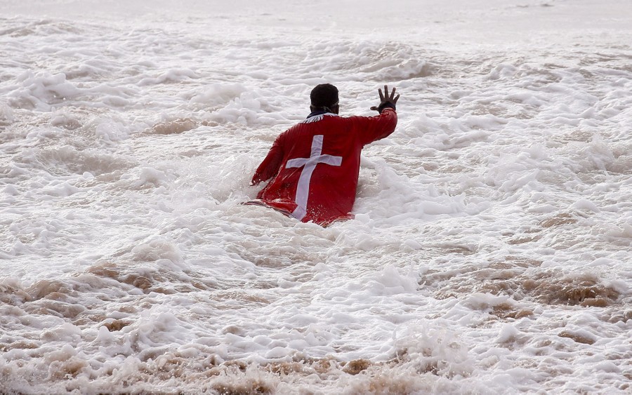 A person prays while standing in in churning water near a shore.