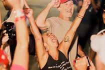 A white woman wearing a black tank top is smiling and throws her arms in the air.