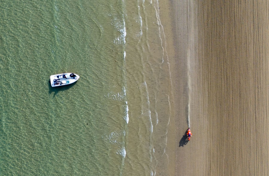 An aerial view of a motocross rider racing along a beach, with a small boat just offshore