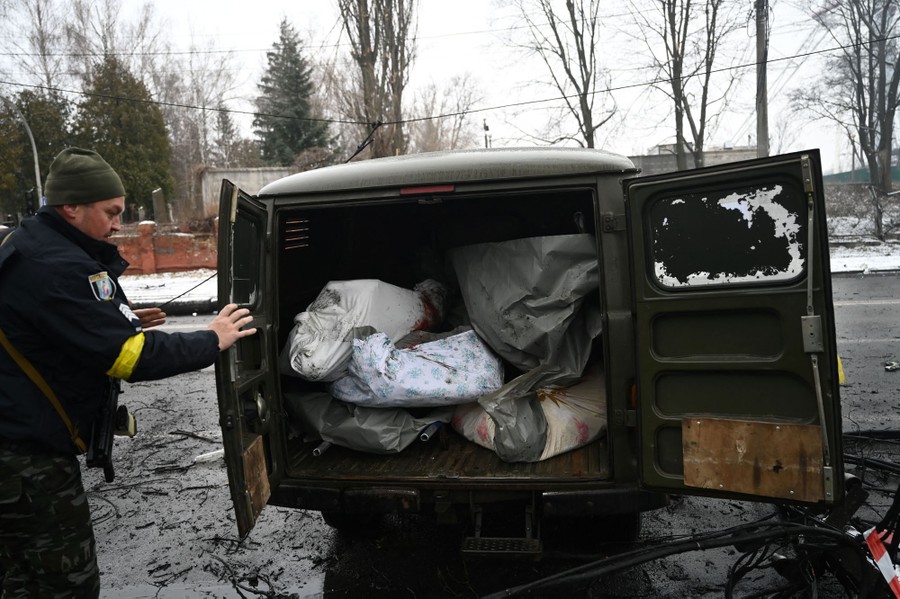 A police officer closes the back door of a truck. Inside can be seen at least five bodies wrapped in sheets or tarps.