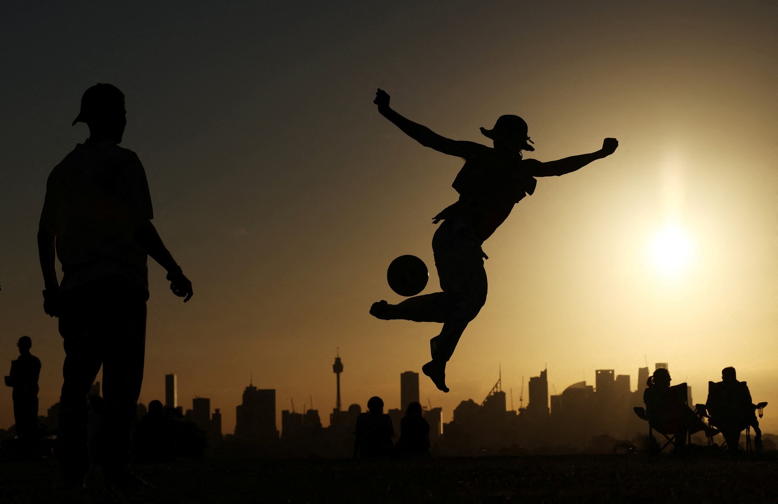 People play, kicking a ball, seen in silhouette, with a city skyline in the background.