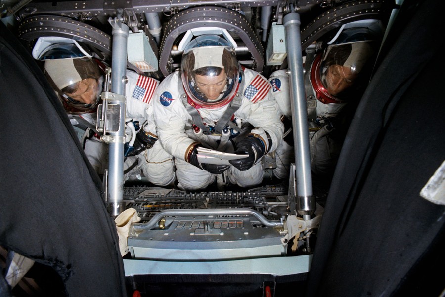 Three astronauts sit side-by-side inside a spacecraft simulator.