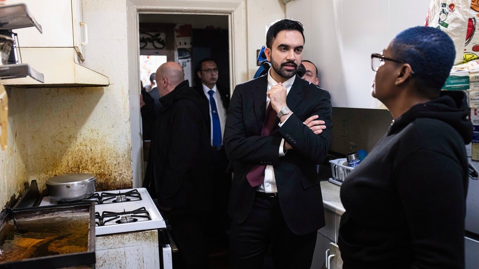 Color photograph of Zohran Mamdani in an apartment with a tenant, standing beside a rusty stovetop.