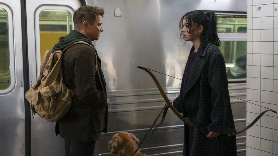 Clint and Kate look at each other while standing on a subway platform. Clint wears a backpack and Kate holds a bow.