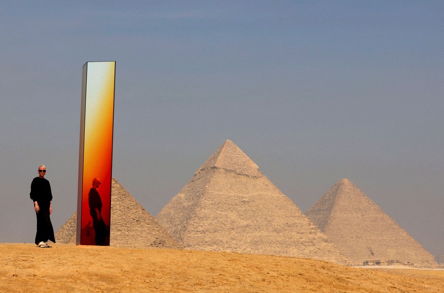 A person stands beside a tall reflective art installation, set in front of the Pyramids of Giza.
