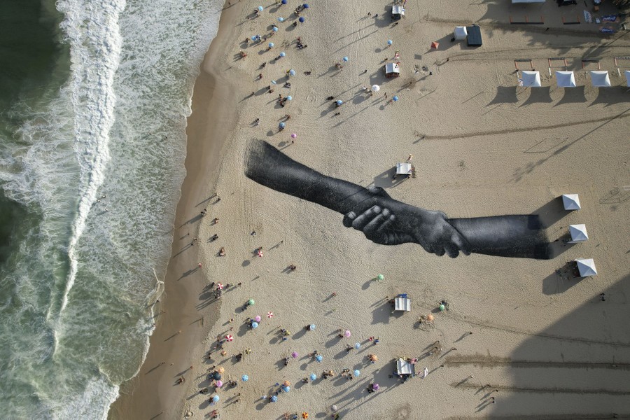 An aerial view of a beach with a large painting on the sand depicting two arms grasping each other.