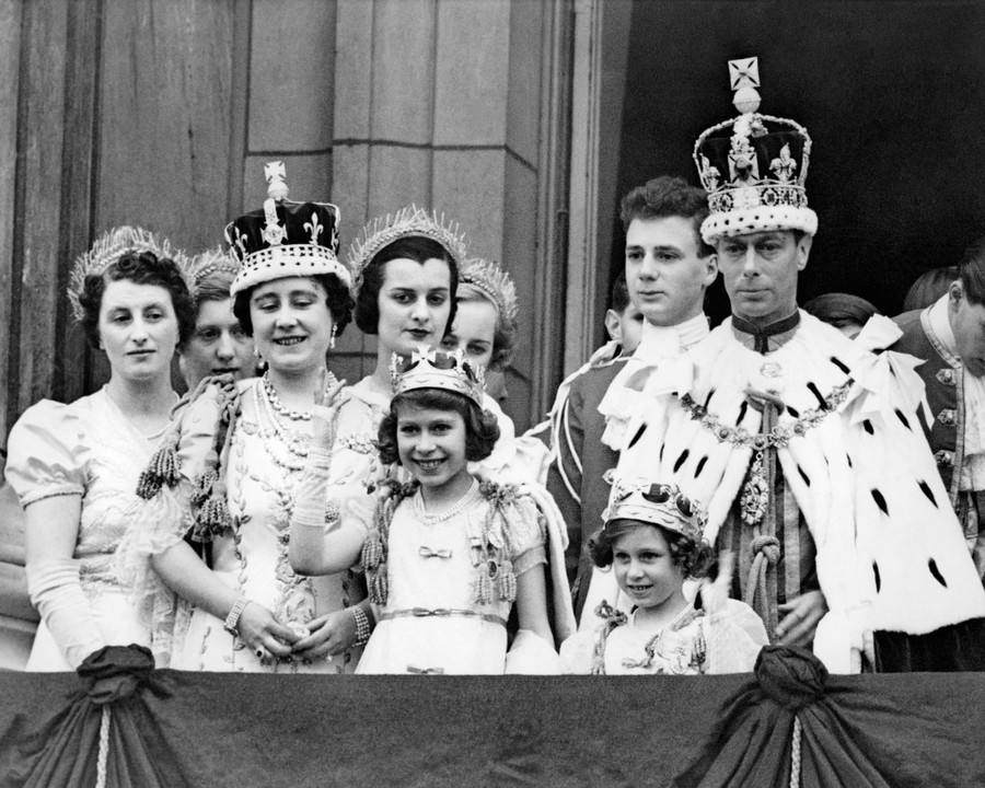 Members of the Royal Family, wearing crowns, gather on a balcony. Young Elizabeth smiles and waves.