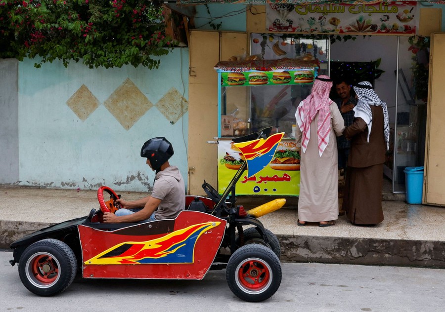 A young man drives a low hand-built car beside a food cart.