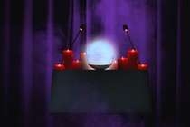 A speaker's lectern in a dark room, against a purple curtain, is crowded with candles and a glowing crystal ball.