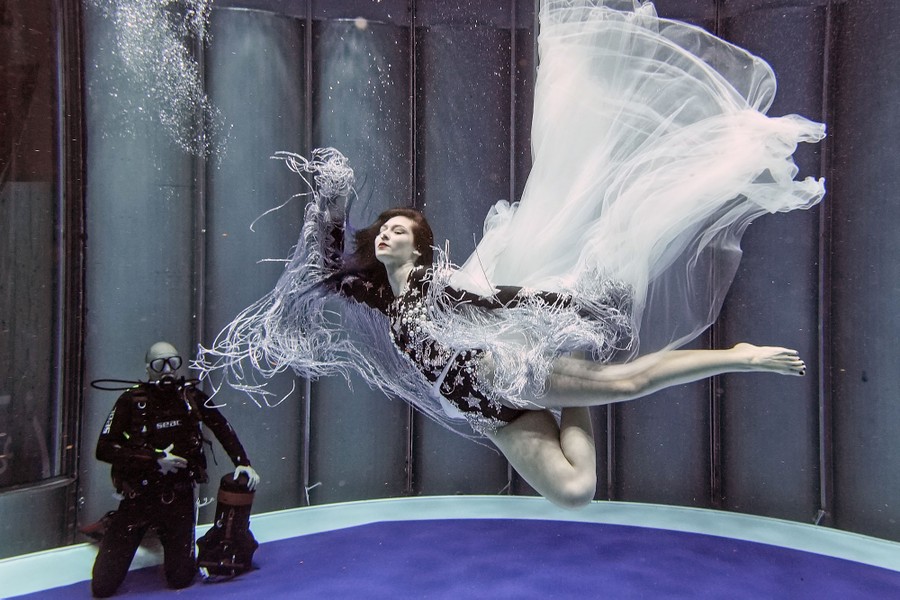 A diver watches a costumed woman free dive in a water tank.