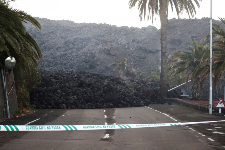 Dark volcanic rocks sit piled up in a flow that covers a road and several palm trees.