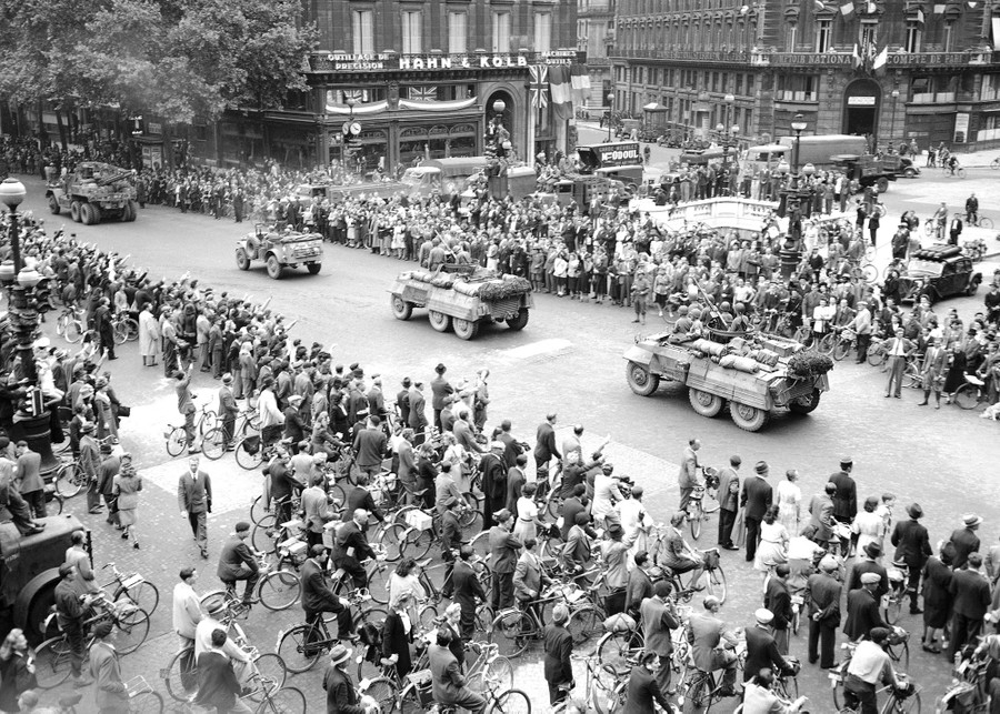 A crowd of French civilians, many on bikes, watch a parade of U.S. soldiers in vehicles pass by.
