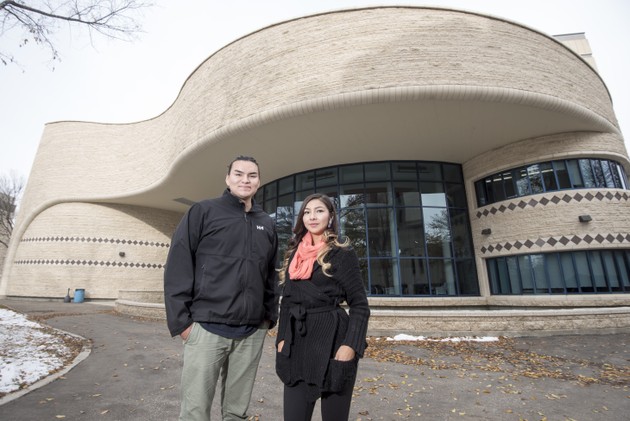 Two students pose in front of a building. There is a thin layer of snow on the ground. 