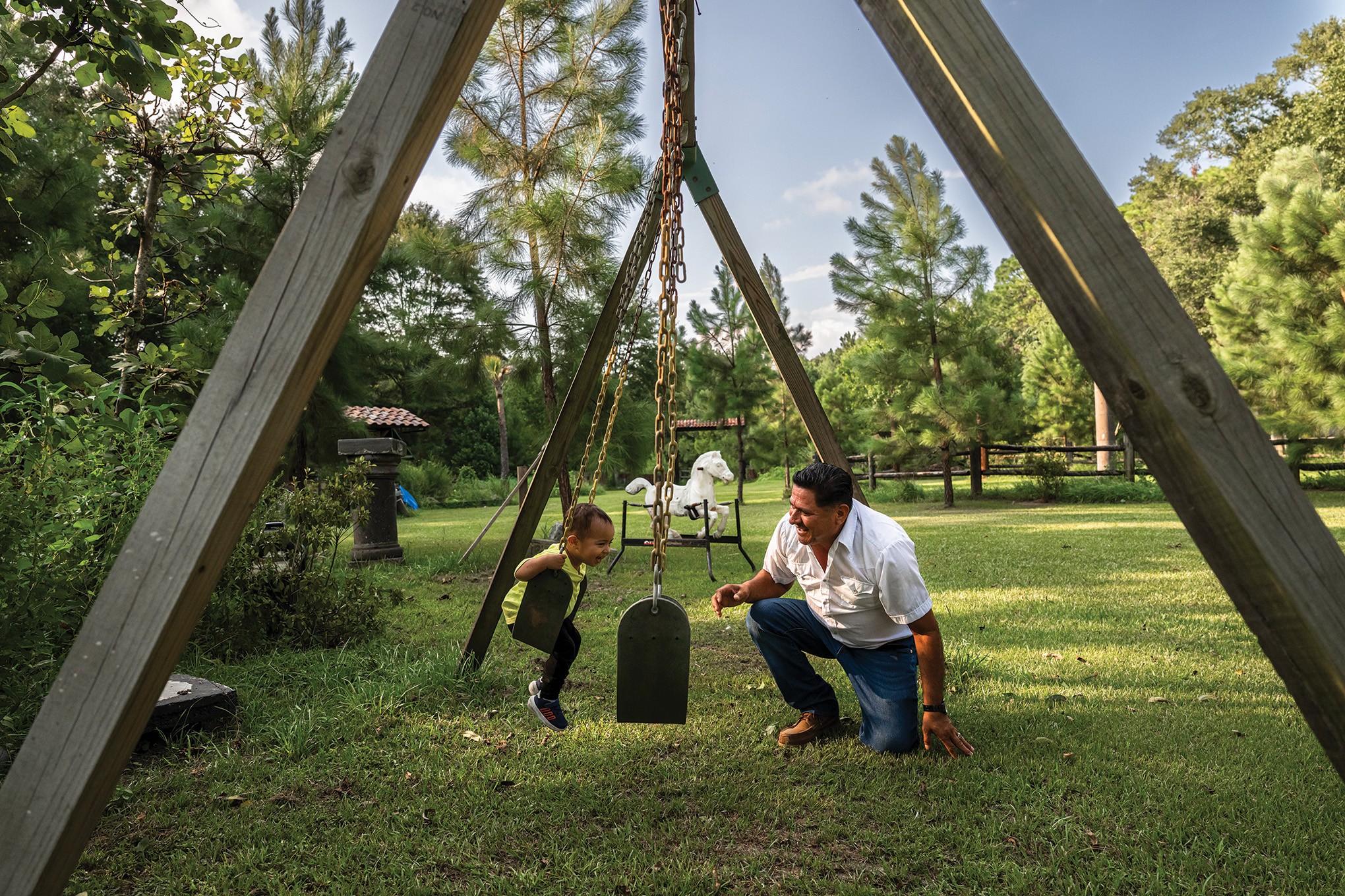 photo of man kneeling on ground with toddler swinging on large swing-set in yard with green grass and trees