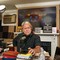 Photo of Steve Bannon sitting in cluttered home studio at microphone on desk covered with books near lighting equipment, in background a TV with still from the movie "Turning Red" over fireplace mantel with signs