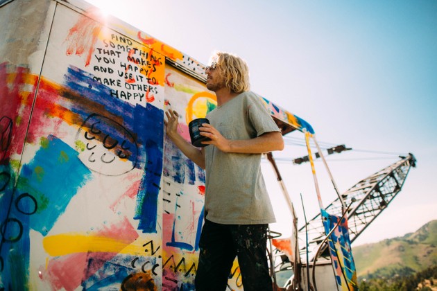 A man stands next to a colorful crane with the words "Find something that makes you happy and use it to make others happy" painted on it.