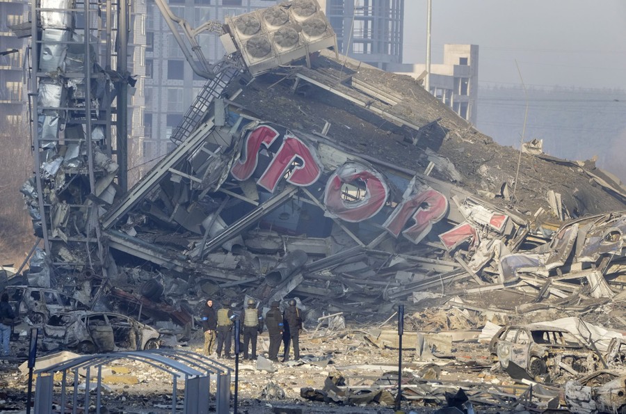 Um pequeno grupo de pessoas está ao lado dos escombros de um prédio destruído, parte de um shopping center, com a inscrição "SPORT" visível nos destroços.