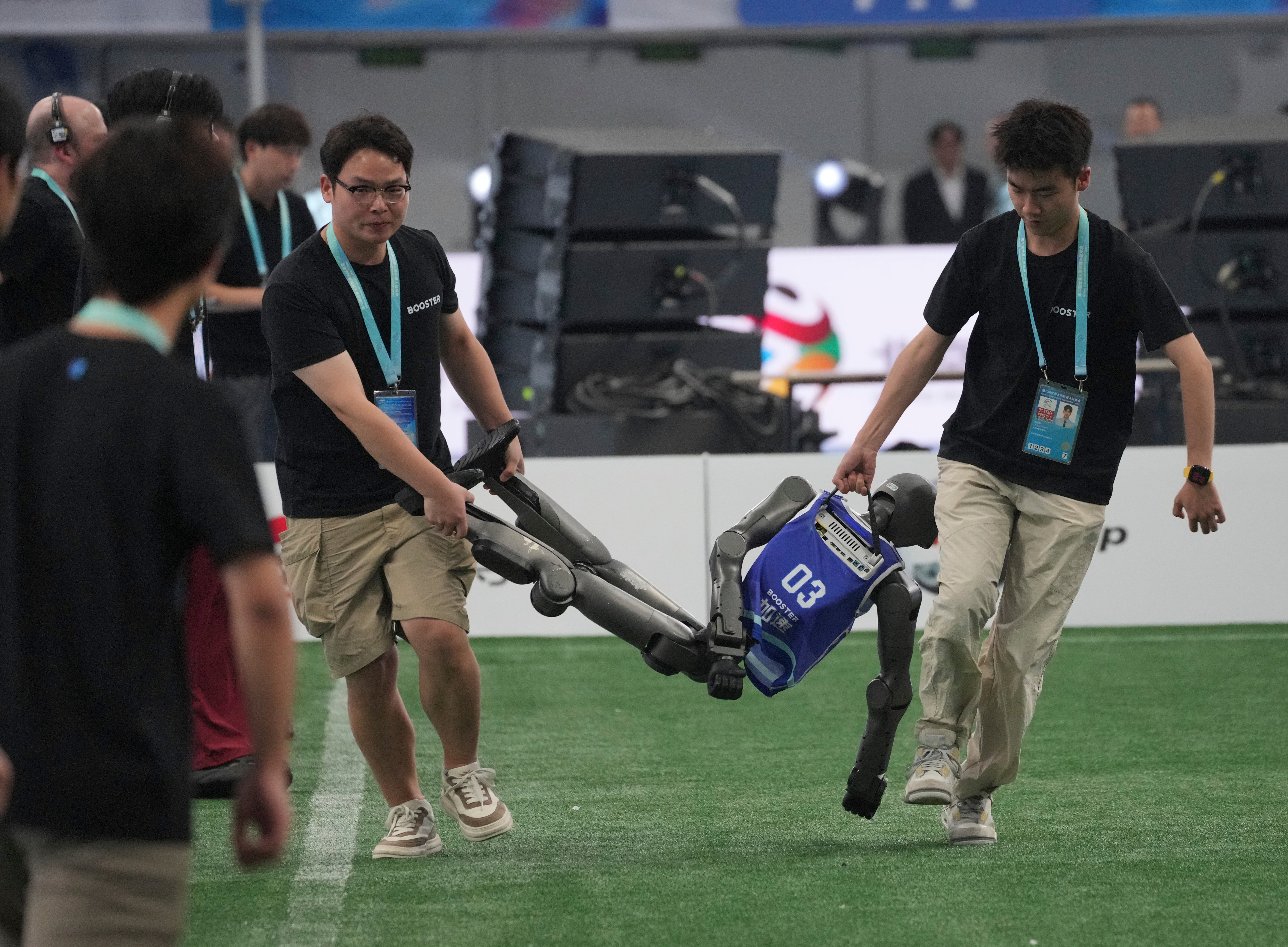 Two people carry an inert humanoid robot off a soccer field.