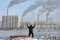 A photograph of a person facing away from the camera, standing with their arms raised, in front of factories with smokestacks emitting smoke