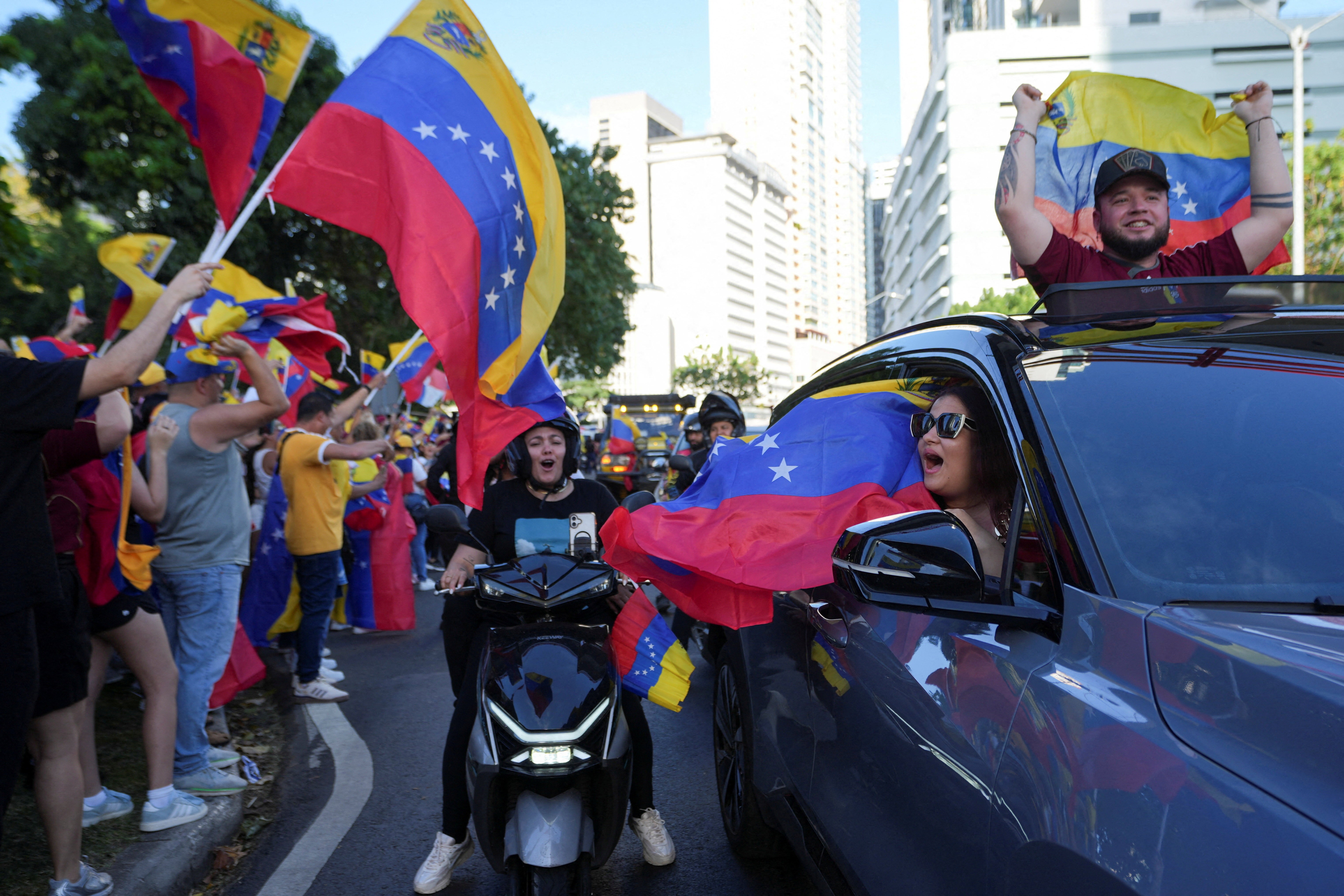 A group of people waving Venezuelan flags greet others driving past who cheer and wave their own flags.