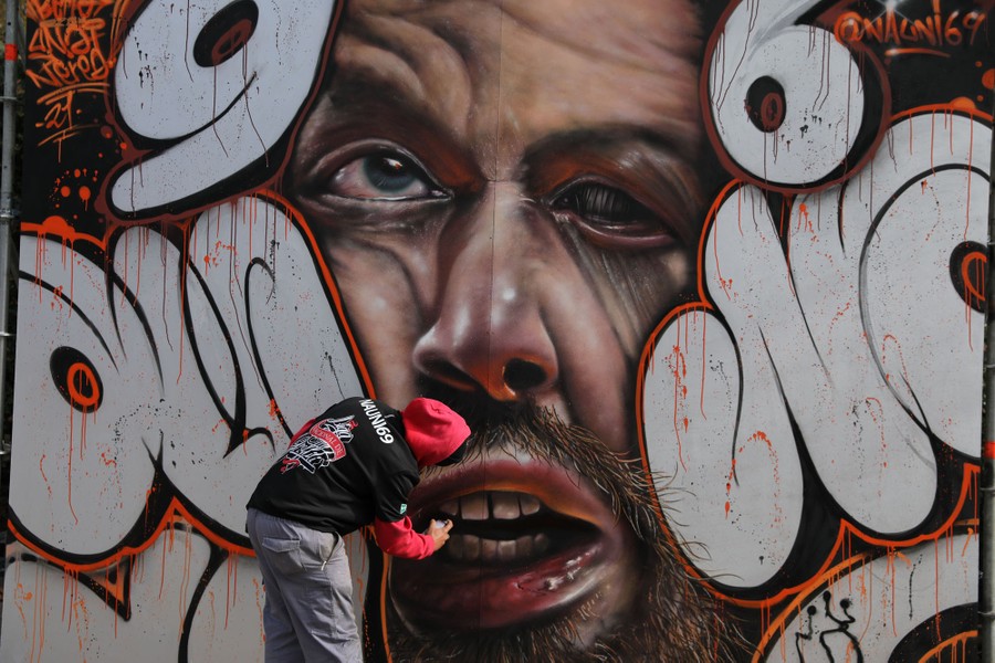 A graffiti artist works on a large mural depicting a person's squished face.